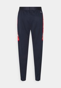 Námořnické modré sportovní kalhoty s červenými vzorovanými bočními panely, elastickým pasem, zúženými nohavicemi a zipy na kotnících, zobrazené ze zadní strany.