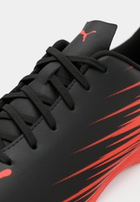 Musta urheilukenkä, jossa punaisia vinoraitoja, teksturoitu synteettinen päällinen, tasaiset mustat nauhat ja logo päällä. Matalaprofiilinen muotoilu.