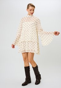 Kreemi mekko, jossa on mustia pilkkuja, kellotetuilla hihoilla, kerroksellisella rypytyksellä ja korkealla kauluksella. Yhdistetty ruskeisiin polven yli ulottuviin saappaisiin.