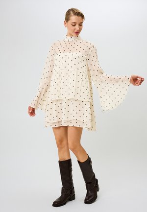 Robe crème à pois noirs, manches cloche, design à volants superposés et col montant. Associée à des bottes brunes jusqu'au genou.