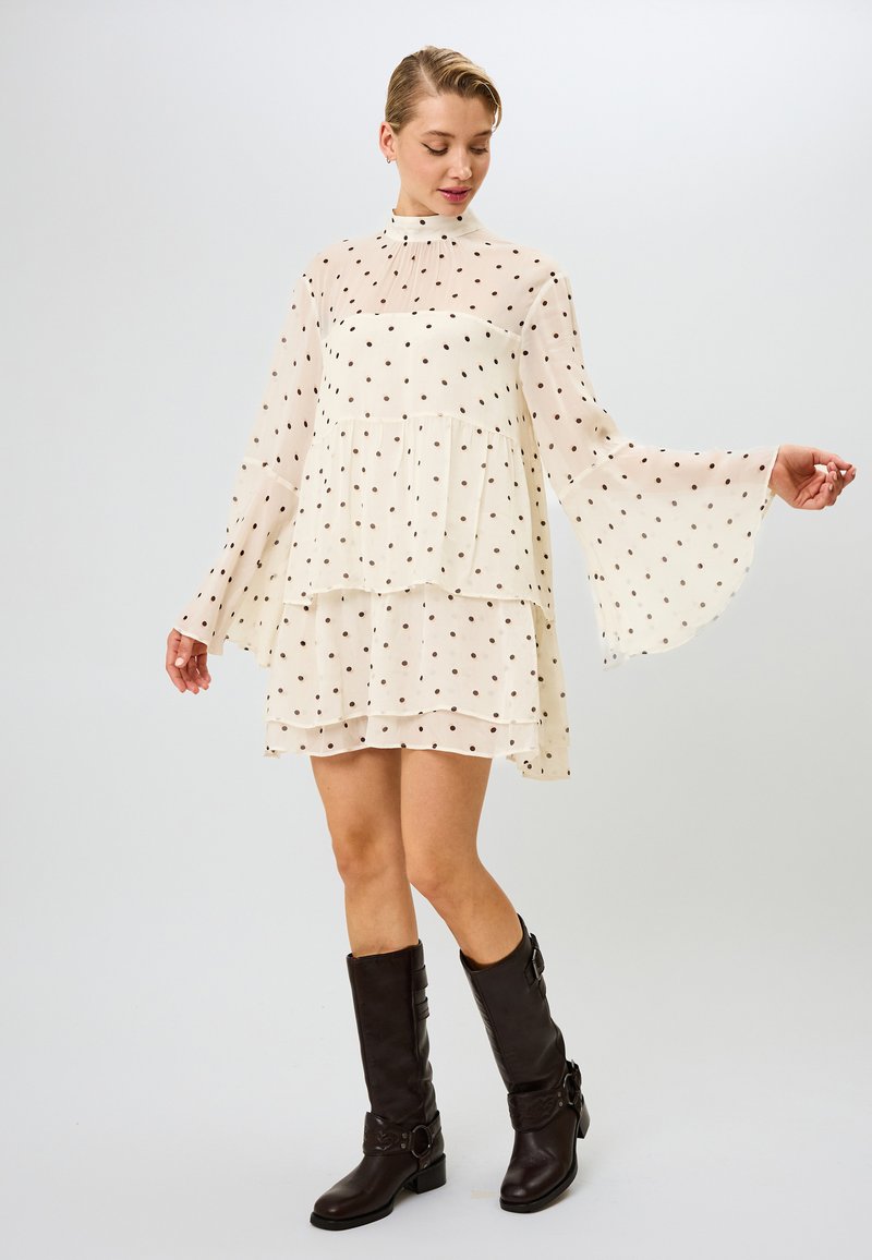 Kreemi mekko, jossa on mustia pilkkuja, kellotetuilla hihoilla, kerroksellisella rypytyksellä ja korkealla kauluksella. Yhdistetty ruskeisiin polven yli ulottuviin saappaisiin.