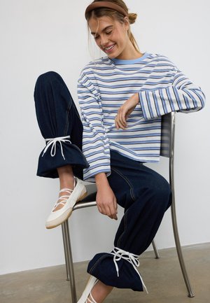 Femme souriante assise sur une chaise en métal, portant un chemisier à rayures bleues et blanches à manches longues, un jean foncé avec des ourlets noués, et des ballerines blanches.