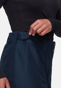 Mörkblå byxor med kardborrestängning i midjan, som har en slät yta och silverfärgade knappdetaljer. Närbild av spännområdet.