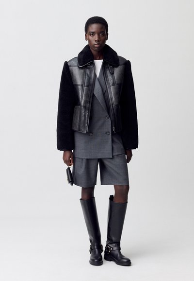 Chaqueta de cuero negro con forro de piel de oveja, pantalones cortos grises a medida, botas negras hasta la rodilla con herrajes, llevando un pequeño bolso negro.