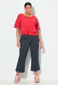 Top corto rojo de manga corta con nudo al frente, pantalones de mezclilla oscuros de pierna ancha y zapatillas rojas con rayas blancas. Tejido suave, corte relajado y diseño único.