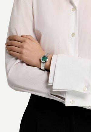 Montre en argent avec un cadran rond vert, sur un bracelet gris clair, portée sur une chemise blanche à manches longues avec des boutons.