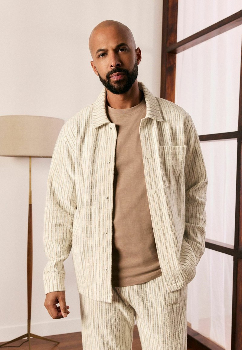 Cream colored shirt jacket with vertical stripes, long sleeves, and collar, paired with a beige round-neck t-shirt and matching striped pants.
