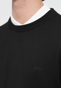 Mężczyzna w czarnym swetrze z okrągłym dekoltem, z subtelnym logo „BOSS” nad kołnierzykiem białej koszuli, zbliżenie na górną część klatki piersiowej i szyję.