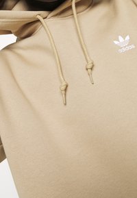 Béžová mikina Adidas s tkaničkami uvázanými do uzlů a bílým logem Adidas na levém hrudníku na světlém pozadí.