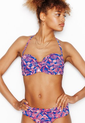 Donna che indossa un bikini blu con motivi di foglie tropicali rosa e bianche, guarda di lato, mani sui fianchi.