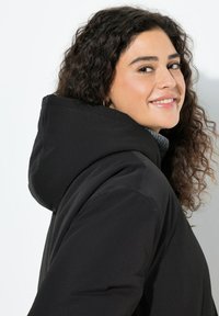 Sort hættejakke med tekstureret stof, tætsiddende design og høj krave, vist mod en lys baggrund.