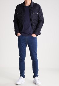 Gequilted marineblauwe jas met drukknoopzakken, marineblauw T-shirt, blauwe jeans en witte sneakers. Focus op texturen en kleurcontrasten in de outfit.