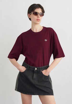 Person iført solbriller, bordeaux Adidas t-shirt og sort denimnederdel med hænder i lommerne mod en ensartet lys baggrund.