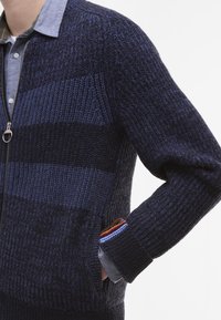 Marine gebreide cardigan met verschillende streeppatronen, ritssluiting en textuurstof. Inclusief contrasterende manchetten in rood, blauw en bruin.