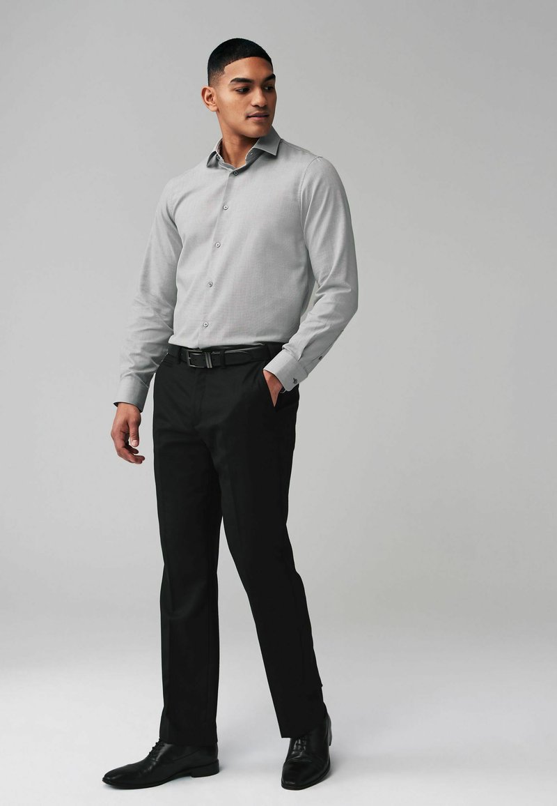 Camicia button-up grigia chiara con un motivo sottile, abbinata a pantaloni neri eleganti e scarpe nere. La camicia presenta un colletto a punte e maniche lunghe.