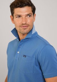 Niebieska koszulka polo wykonana z bawełny, z paskowanym wewnętrznym kołnierzykiem, trzema guzikami z przodu oraz kieszonką z metką marki.