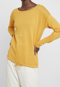 Žlutý dlouhý rukáv top z měkké, strukturované tkaniny. Má široký výstřih a nastavitelné stahovací dno. Stylizováno s bílými kalhotami.