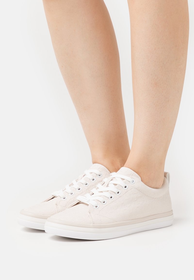 Baskets en toile beige clair avec des lacets blancs portées pieds nus sur un fond blanc uni.