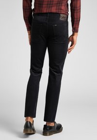 Tmavě modré manšestrové kalhoty slim fit s kapsami na zadní straně a značkovou hnědou koženou nášivkou na pásku. Černé boty doplňují vzhled.