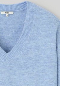 Pull léger bleu en tissu doux, avec un décolleté en V et des poignets côtelés, présentant une texture lisse et des variations de couleur subtiles.
