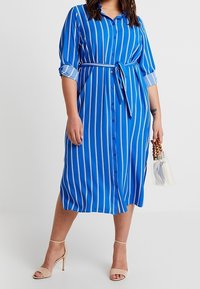 Abito camicia blu con strisce verticali bianche, cintura in vita, maniche 3/4 e vestibilità ampia. Abbinato a una borsa trasparente e tacchi beige.