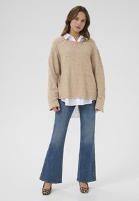Beige Strickpullover mit lockerer Passform, Rundhalsausschnitt und langen Ärmeln, über einem weißen Hemd mit Kragen getragen. Kombiniert mit weit geschnittenen blauen Jeans.