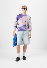Maglione lavorato a maglia con motivi floreali viola e rosa, pantaloni corti in denim azzurro, mocassini neri, calzini bianchi alla caviglia, mentre tiene una borsa a motivi blu.