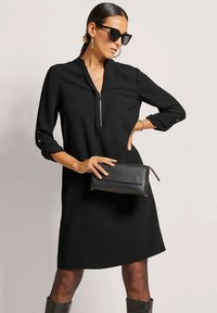 Schwarzes Kleid mit Kragen und einem Reißverschluss auf der Vorderseite, mit langen Ärmeln und abgerundetem Saum. Das Model hält eine schwarze Clutch und trägt Sonnenbrillen.