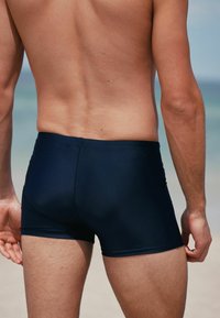 Marine-colored Schwimmshorts mit enganliegendem Schnitt, glatter Textur und dezenten Nähten, von hinten vor einem Strandhintergrund betrachtet.