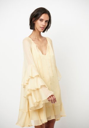 Vrouw draagt een lichtgele, luchtige jurk met gelaagde, geplooide mouwen, staand tegen een effen witte achtergrond, licht naar links gewend.