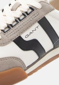 Αθλητικό παπούτσ ι με μπεζ νάιλον σώμα, γκρι σουέντ λεπτομέρειες και μαύρη δερμάτινη ρίγα. Οι κορδόνια είναι λευκά· η σόλα είναι καφέ από καουτσούκ. Εμφανίζεται το λογότυπο της μάρκας.