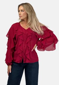 Rote Chiffonbluse mit Rüschen entlang der Vorderseite und den Ärmeln, V-Ausschnitt und Knopfverschluss. Kombiniert mit dunklen Jeans.
