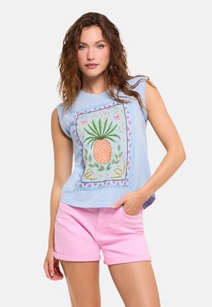 Joven mujer con cabello rizado castaño, vestida con una camiseta azul sin mangas con bordado de piña y pantalones cortos rosas, de pie con la mano en el bolsillo.
