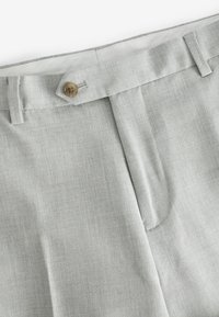 Pantalones gris claro con una pestaña de cinturilla abotonada y trabillas, mostrando un primer plano del cierre frontal y la textura de la tela.