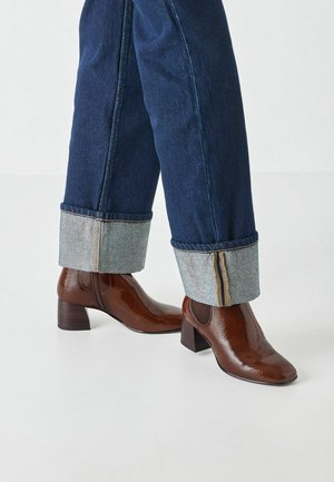 Bottines marron brillantes avec un talon empilé ; associées à un jean bleu foncé avec des poignets retroussés en denim clair contrastant.