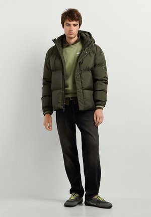 Jeune homme debout portant une doudoune vert foncé sur un sweat olive, un jean noir et des baskets grises avec des lacets jaunes sur fond blanc.
