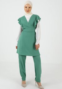 Vihreä tunika, jossa on frillat, yhdistettynä samanvärisiin housuihin. Valmistettu teksturoidusta kankaasta, V-päällekkäisrakenteella, valkoisen paidan päällä.