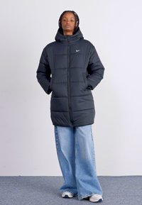 Must välja voodriga must jope, millel on kapuuts, esipaneelil lukuga ja küljetaskud. Sile tekstuur, rinnal brändi logo, kantud laia jalgadega teksade peal.