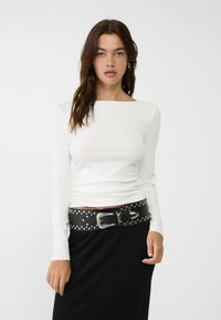 White long-sleeve top with a boat neckline, paired with a black studded belt featuring a silver buckle, worn over a black skirt.