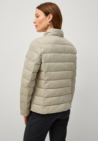 Chaqueta acolchada en beige claro, fabricada con un material suave y brillante. Cuenta con un cuello alto y costuras horizontales. Combinada con pantalones negros.