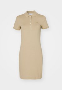 Robe polo beige avec un col, des manches courtes et une patte de boutonnage. Présente un discret logo brodé sur la poitrine. Texture de tissu lisse.