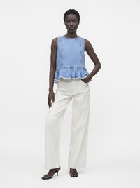 Femme portant un top en denim bleu sans manches à basque, un pantalon blanc à jambes larges et des sandales à talons noires, debout devant un fond clair uni.
