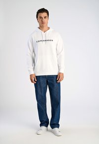 Felpa bianca con la scritta "COPENHAGEN", tasca a marsupio frontale e cappuccio con cordino. Abbinata a jeans azzurri a gamba dritta e sneakers bianche.