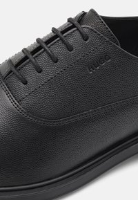 Chaussures en cuir noir présentant une texture granuleuse, un bout rond et un design classique à lacets. Agrémentées d'un logo de marque discret.