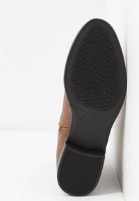 Chaussure en cuir marron avec une finition lisse, dotée d'une semelle en caoutchouc noir à texture ondulée et d'un petit indicateur de taille sur le côté.