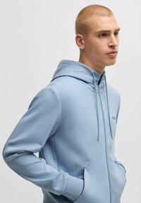 Ljusblå zip-up hoodie av slät tyg. Har en huva med snörning, framfickor och en minimalistisk logotyp på bröstet.
