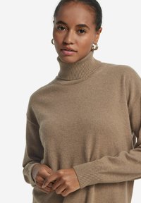 Beige Rollkragenpullover aus weichem, strukturiertem Material. Mit geripptem Kragen und Bündchen, in entspanntem Schnitt. Nahaufnahme von Händen zu sehen.