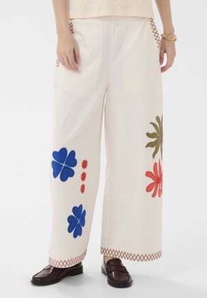 Witte wijd uitlopende broek met blauwe, rode en olijfgroene bloemrijke borduursels, kruissteekdetails bij de zakken en zomen, gedragen met donkere loafers.