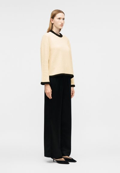 Femme portant un pull crème avec bordure noire, pantalon large noir et chaussures noires à talons bas et pointues, debout devant un fond uni.