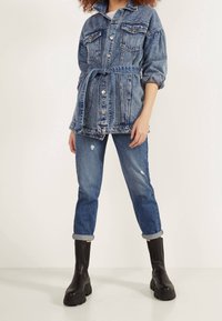 Blue denim jacket with a belted waist, featuring two chest pockets and button closures. Paired with distressed blue jeans and black platform boots.